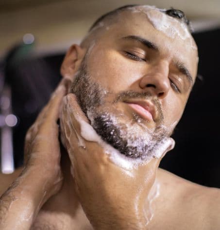 Person washing face in shower to maintain beard hygiene.