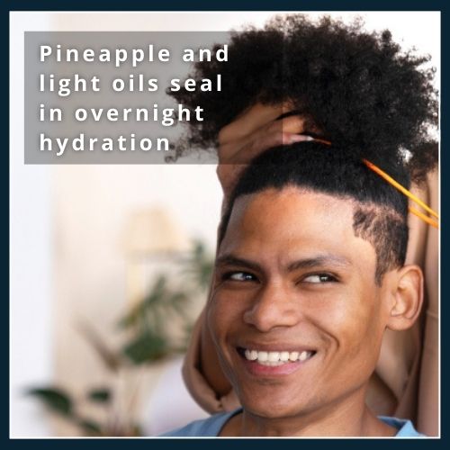 Man with curls piled in a soft scrunchie applying a few drops of lightweight oil before sleep, illustrating the pineapple method and light oils to lock in overnight moisture.