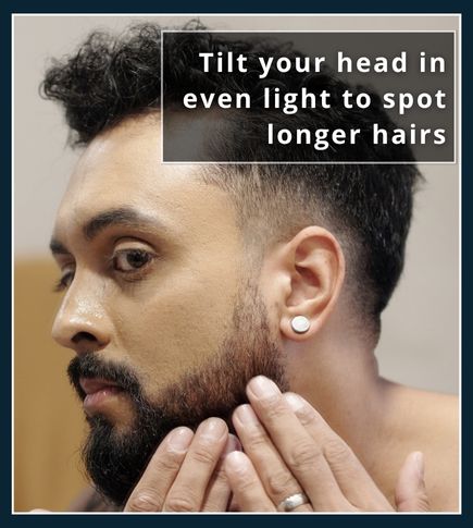 Man tilting his head in front of a well‑lit mirror to inspect uneven beard hairs