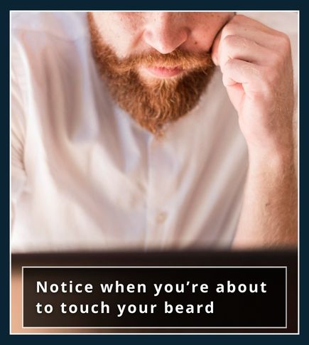 A man sitting at a desk with his hand halfway to his beard, looking thoughtful, showing the habit of touching.