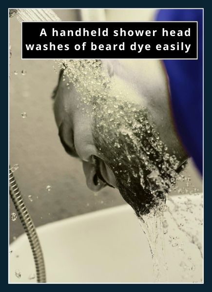 bearded man washing beard dye from his beard in the tub with a handheld shower head pouring water over his beard