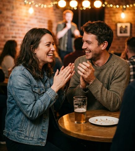 A man and a woman sitting close together at a small table during an open mic or comedy night. They’re laughing and clapping, with warm stage lights and a blurred performer holding a mic in the background