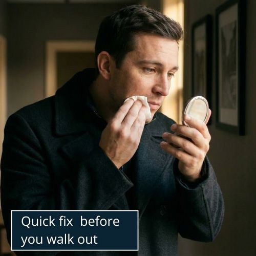 a well-groomed man standing in his hallway, gently wiping the corner of his mouth with a folded tissue while holding a small compact mirror slightly to the side.