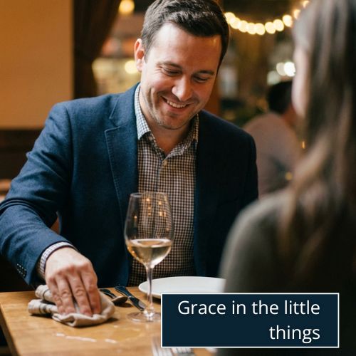 A man on a dinner date in a softly lit restaurant, casually wiping a small water spill on the table with a napkin while smiling.