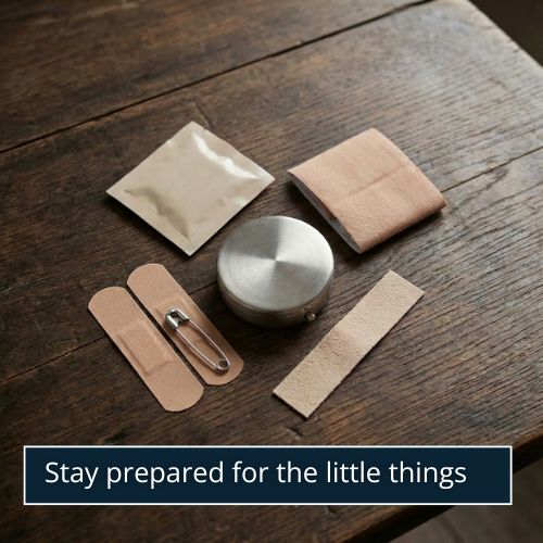 A flat lay of small men’s essentials on a dark wooden surface: a stain remover wipe packet, a silver safety pin, a small moleskin strip, two plasters, a compact pill case, and a bit of folded fashion tape.
