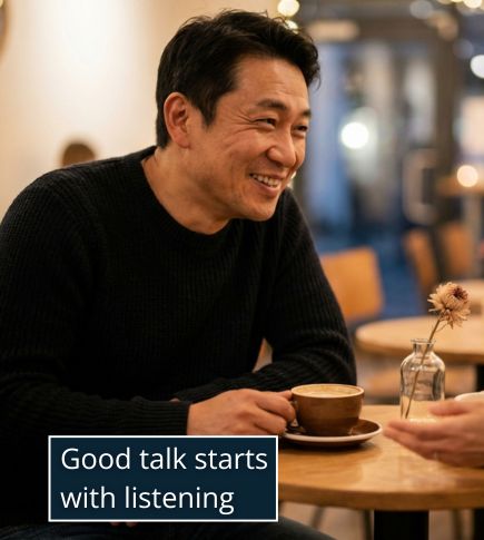 A man and woman sitting at a small café table during a date, mid-conversation. The man is smiling and listening attentively with relaxed posture, one hand resting near a coffee cup.