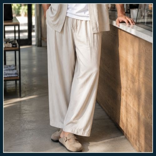Man waiting at counter in coffee barm he is wearing relaxed kimono style overshirt, oversize tshirt and oversized wide leg linen trousers with suede clogs