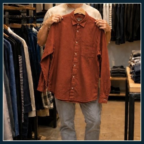 Man in clothing store holding up a light coloured top to his body.